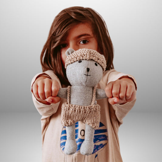 Child holding mini plush teddy bear with knitted hat and overalls against neutral background – Mamutino